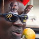 A portrait of a man wearing black-lens sunglasses with a navy frame. On top of the glasses are two paper cutouts of Raila Odinga. On the frames are more paper cutouts including a large, orangey-yellow letter "R" and the words "Raile Amolo Odinga." Around the man's neck appears to be some kind of giant faux-fruit (mango?) necklace. He is also wearing a red helmet.